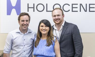 The image shows three people standing together in front of a sign that reads "HOLOCENE." The person on the left is a man with short brown hair, wearing a light blue dress shirt. The person in the middle is a woman with long dark hair, wearing a sleeveless blue top. The person on the right is a man with short blonde hair, a beard, and is dressed in a dark grey suit with a white shirt underneath. All three individuals are smiling and appear to be posing for the photo.