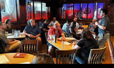 A group of people gathered in a casual restaurant setting, seated around several tables while attentively listening to a speaker standing to the right. The speaker is wearing a gray shirt and khaki pants, gesturing with both hands as they address the audience. The atmosphere is relaxed, with a variety of drinks and menus on the tables, and the backdrop features large, illuminated beer tanks behind glass windows. The overall mood appears engaged and informal, suggesting a collaborative or networking event.