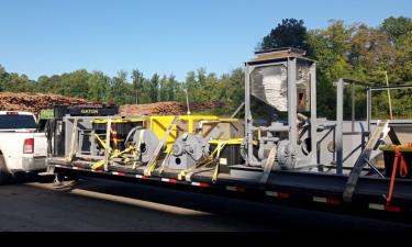 A large industrial air quality system is loaded onto a flatbed trailer, secured with yellow straps. The equipment includes a series of motors, fans, and ducts, with wood piles and green trees visible in the background, indicating an outdoor setting. The equipment appears to be part of an emissions control or filtration unit, ready for transport.