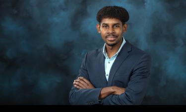 A professional portrait of a man with medium skin tone, short curly hair, and a confident smile. He is dressed in a dark navy blazer over a light blue collared shirt. His arms are crossed in a relaxed pose. The background is a textured dark blue, adding a formal and professional feel to the image.