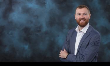  A professional portrait of a man with a beard and short, reddish-brown hair. He is wearing a light blue blazer over a white button-up shirt. The man is smiling confidently with his arms crossed. The background is a soft, textured mix of dark blue and gray tones, giving the image a polished and formal look.