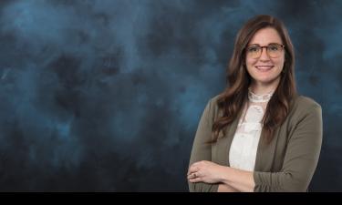 Portrait of a woman with long, wavy brown hair, wearing glasses with a patterned frame. She is dressed in a light lace blouse under a green blazer and is smiling with her arms crossed. The background is a dark, textured gradient with shades of blue and black.