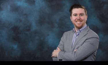 Portrait of a man with a friendly smile, wearing a light gray blazer and a blue striped shirt, standing with arms crossed in front of a dark blue, cloud-like background.