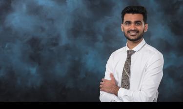 Portrait of Vinit Chaudhary, a Cohort 2024 fellow at Innovation Crossroads, smiling with arms crossed. He is wearing a white dress shirt and patterned tie against a dark blue textured background.