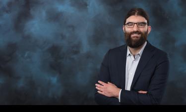 Professional portrait of a man with long dark hair and a beard, wearing glasses and a dark suit jacket over a light button-up shirt. He stands with his arms crossed, smiling slightly against a textured dark blue background.