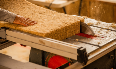 Close-up of a hand guiding a large, thick block of natural insulation material through a table saw, producing fine particles as it is cut. The block has a fibrous, organic texture, suggesting it's made from sustainable materials