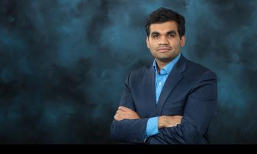 A professional headshot of a man wearing a dark blue suit and light blue shirt. He has his arms crossed and is looking confidently at the camera. The background is a textured, dark teal-blue color.