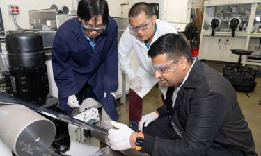 Three researchers wearing safety goggles and lab coats collaborate in a laboratory, closely examining a high-tech piece of equipment with rollers and metallic components.