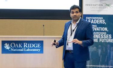 Manas Pathak, an Innovation Crossroads Cohort 2023 fellow, presenting at Oak Ridge National Laboratory. He is standing behind a podium with the Oak Ridge National Laboratory logo, delivering a talk with a microphone in hand. A banner in the background highlights the Innovation Crossroads program's mission to empower leaders, drive innovation, and launch businesses for the future.