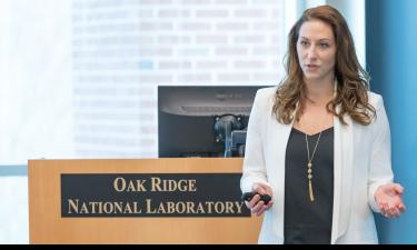 Megan O'Connor delivering a presentation at Oak Ridge National Laboratory, standing behind a podium with the lab's logo. She is wearing a white blazer and black top, gesturing with one hand while holding a presentation remote in the other.