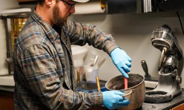 A man with blue gloves mixes materials in a laboratory setting, focused on creating a new product. The background includes shelves with equipment and tools related to his work. His expression is intent as he works with precision, contributing to his innovative efforts in recycling glass.