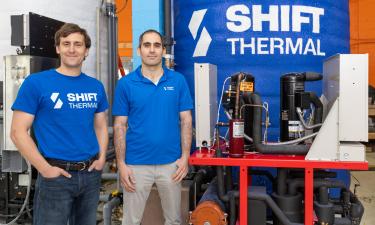 Two men stand side by side in an industrial setting, both wearing blue "SHIFT THERMAL" shirts. The man on the left wears a t-shirt and jeans, while the man on the right wears a collared shirt and light-colored pants. Behind them is a large blue thermal storage unit with the "SHIFT THERMAL" logo printed on it. To their right, a red industrial refrigeration or thermal management system with black compressors, pipes, and wiring is visible. The background features various pipes, equipment, and an orange-painted