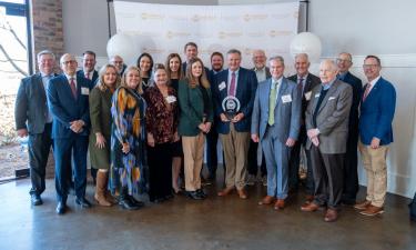 A group of professionally dressed people is gathered in an indoor event space, posing for a photo in front of a backdrop with the Knoxville Chamber and Pinnacle Business Awards logos. The central figure holds a circular award plaque, and everyone is smiling. The setting has natural light coming in from a window on the left, with a brick wall visible. White balloons are present, adding to the celebratory atmosphere.