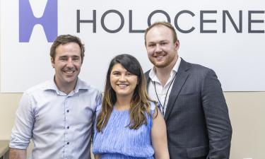Three people stand smiling in front of a wall with the Holocene logo. The group includes two men—one in a light button-down shirt, the other in a dark blazer—and a woman in a sleeveless blue dress, standing between them. They appear to be part of a team or event celebrating a milestone.