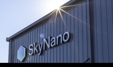 Exterior view of a metal building with the SkyNano logo mounted on the side. The sun shines brightly over the roof, creating a lens flare and casting light rays across the image.