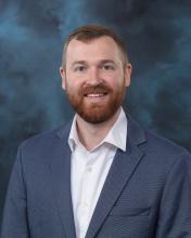 A professional headshot of a man smiling and looking directly at the camera, wearing business attire, with a neutral blue backdrop.