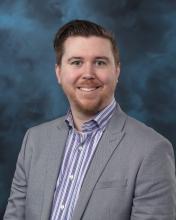 A professional headshot of a man smiling and looking directly at the camera, wearing business attire, with a neutral blue backdrop.