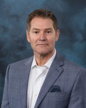 A professional headshot of a man smiling and looking directly at the camera, wearing business attire, with a neutral blue backdrop.