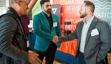 Three men wearing name tags are engaged in conversation at a professional event. Two of them are shaking hands, while the third stands nearby smiling. They are in front of a colorful backdrop with text that includes "Berkeley Commons" and "Future."
