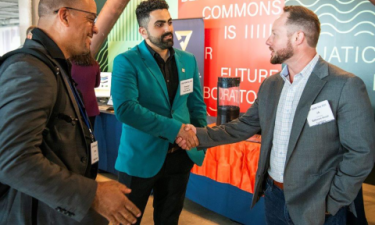 Three men wearing name tags are engaged in conversation at a professional event. Two of them are shaking hands, while the third stands nearby smiling. They are in front of a colorful backdrop with text that includes "Berkeley Commons" and "Future."