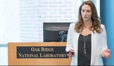 Megan O'Connor delivering a presentation at Oak Ridge National Laboratory, standing behind a podium with the lab's logo. She is wearing a white blazer and black top, gesturing with one hand while holding a presentation remote in the other.