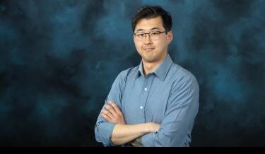 A professional portrait of a man with short black hair, glasses, and a light blue button-up shirt. He stands with his arms crossed against a dark blue textured background, looking confidently at the camera.