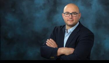 Portrait of a bald man wearing blue-framed glasses, a dark blazer, and a light blue striped shirt, standing with arms crossed against a dark blue studio background.