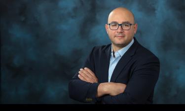 Portrait of a bald man wearing blue-framed glasses, a dark blazer, and a light blue striped shirt, standing with arms crossed against a dark blue studio background.
