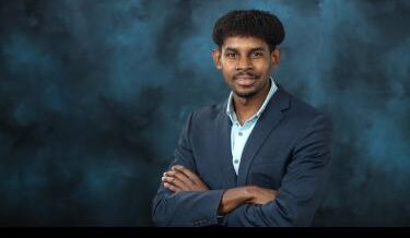 A professional portrait of a man with medium skin tone, short curly hair, and a confident smile. He is dressed in a dark navy blazer over a light blue collared shirt. His arms are crossed in a relaxed pose. The background is a textured dark blue, adding a formal and professional feel to the image.