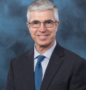 Professional headshot of a middle-aged man with short gray hair, wearing glasses, a dark suit jacket, a blue tie, and a light blue striped shirt, posed in front of a blue studio background.