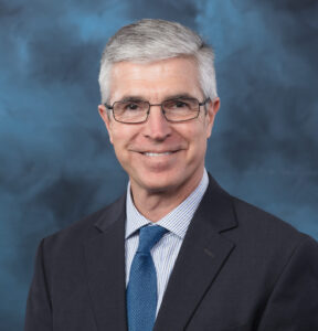 Professional headshot of a middle-aged man with short gray hair, wearing glasses, a dark suit jacket, a blue tie, and a light blue striped shirt, posed in front of a blue studio background.