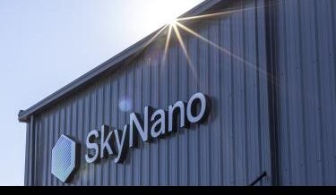 Exterior view of a metal building with the SkyNano logo mounted on the side. The sun shines brightly over the roof, creating a lens flare and casting light rays across the image.