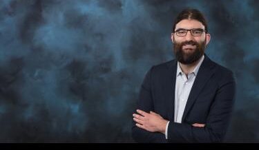 Professional portrait of a man with long dark hair and a beard, wearing glasses and a dark suit jacket over a light button-up shirt. He stands with his arms crossed, smiling slightly against a textured dark blue background.