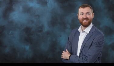  A professional portrait of a man with a beard and short, reddish-brown hair. He is wearing a light blue blazer over a white button-up shirt. The man is smiling confidently with his arms crossed. The background is a soft, textured mix of dark blue and gray tones, giving the image a polished and formal look.