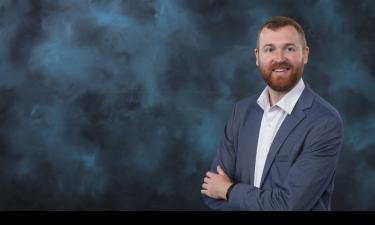 A professional portrait of a man with a beard and short, reddish-brown hair. He is wearing a light blue blazer over a white button-up shirt. The man is smiling confidently with his arms crossed. The background is a soft, textured mix of dark blue and gray tones, giving the image a polished and formal look.