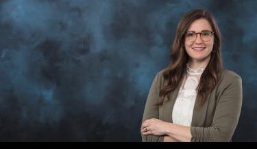 Portrait of a woman with long, wavy brown hair, wearing glasses with a patterned frame. She is dressed in a light lace blouse under a green blazer and is smiling with her arms crossed. The background is a dark, textured gradient with shades of blue and black.