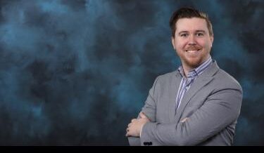 Portrait of a man with a friendly smile, wearing a light gray blazer and a blue striped shirt, standing with arms crossed in front of a dark blue, cloud-like background.