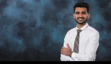 Portrait of Vinit Chaudhary, a Cohort 2024 fellow at Innovation Crossroads, smiling with arms crossed. He is wearing a white dress shirt and patterned tie against a dark blue textured background.