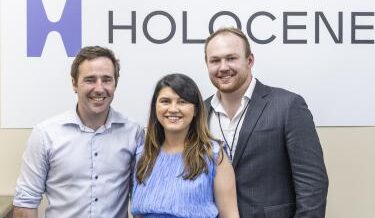 The image shows three people standing together in front of a sign that reads "HOLOCENE." The person on the left is a man with short brown hair, wearing a light blue dress shirt. The person in the middle is a woman with long dark hair, wearing a sleeveless blue top. The person on the right is a man with short blonde hair, a beard, and is dressed in a dark grey suit with a white shirt underneath. All three individuals are smiling and appear to be posing for the photo.