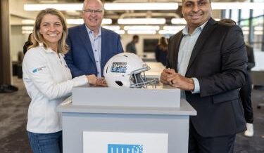 Rajan Kumar, co-founder and CEO of Ateios Systems, stands smiling in a business suit next to two TitletownTech representatives. They are posing around a white podium featuring a football helmet with the "TitletownTech Startup Draft" logo on it, inside a modern, bright office space.