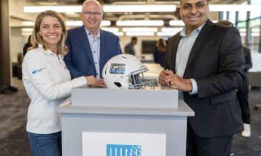 Rajan Kumar, co-founder and CEO of Ateios Systems, stands smiling in a business suit next to two TitletownTech representatives. They are posing around a white podium featuring a football helmet with the "TitletownTech Startup Draft" logo on it, inside a modern, bright office space.
