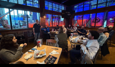 A group of people gathered in a brewery restaurant, listening to a speaker standing near a window with large brewing tanks illuminated by blue and red lights in the background. The audience is seated at tables with food baskets and drinks, attentively engaged in the discussion.