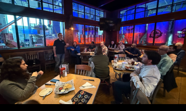 A group of people gathered in a brewery restaurant, listening to a speaker standing near a window with large brewing tanks illuminated by blue and red lights in the background. The audience is seated at tables with food baskets and drinks, attentively engaged in the discussion.
