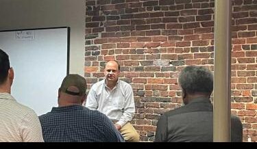 Man sitting on a stool speaking to a group of people in an audience.