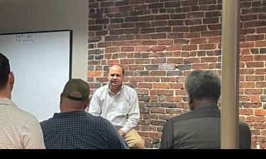 Man sitting on a stool speaking to a group of people in an audience. 
