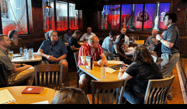 A group of people gathered in a casual restaurant setting, seated around several tables while attentively listening to a speaker standing to the right. The speaker is wearing a gray shirt and khaki pants, gesturing with both hands as they address the audience. The atmosphere is relaxed, with a variety of drinks and menus on the tables, and the backdrop features large, illuminated beer tanks behind glass windows. The overall mood appears engaged and informal, suggesting a collaborative or networking event.