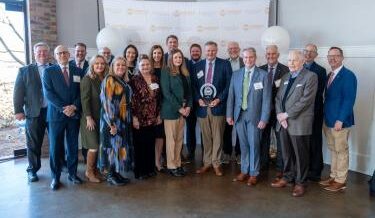 A group of professionally dressed people is gathered in an indoor event space, posing for a photo in front of a backdrop with the Knoxville Chamber and Pinnacle Business Awards logos. The central figure holds a circular award plaque, and everyone is smiling. The setting has natural light coming in from a window on the left, with a brick wall visible. White balloons are present, adding to the celebratory atmosphere.