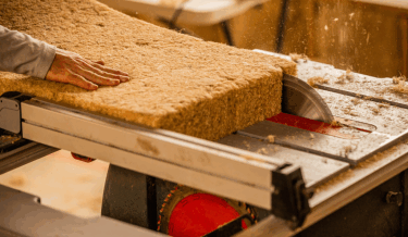 Close-up of a hand guiding a large, thick block of natural insulation material through a table saw, producing fine particles as it is cut. The block has a fibrous, organic texture, suggesting it's made from sustainable materials