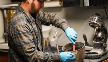 A man with blue gloves mixes materials in a laboratory setting, focused on creating a new product. The background includes shelves with equipment and tools related to his work. His expression is intent as he works with precision, contributing to his innovative efforts in recycling glass.