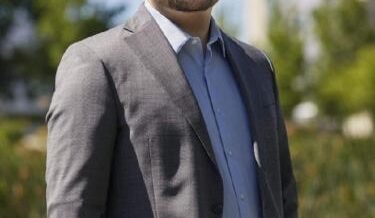 A bald man with a beard and glasses is smiling while standing outdoors. He is wearing a gray suit jacket over a light blue dress shirt. The background features green trees and a blurred modern building under a clear blue sky.